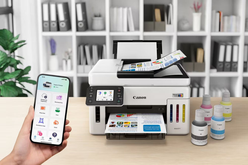 Urządzenie wielofunkcyjne CANON Maxify GX5540 Druk w kolorze, Automatyczny druk dwustronny, Wi-Fi prezentacja urządzenia na blacie obok butelki z atramentem ręka trzymająca telefon oszczędność kosztów częste drukowanie uzupełnienie w prosty i czysty sposób wydajność
