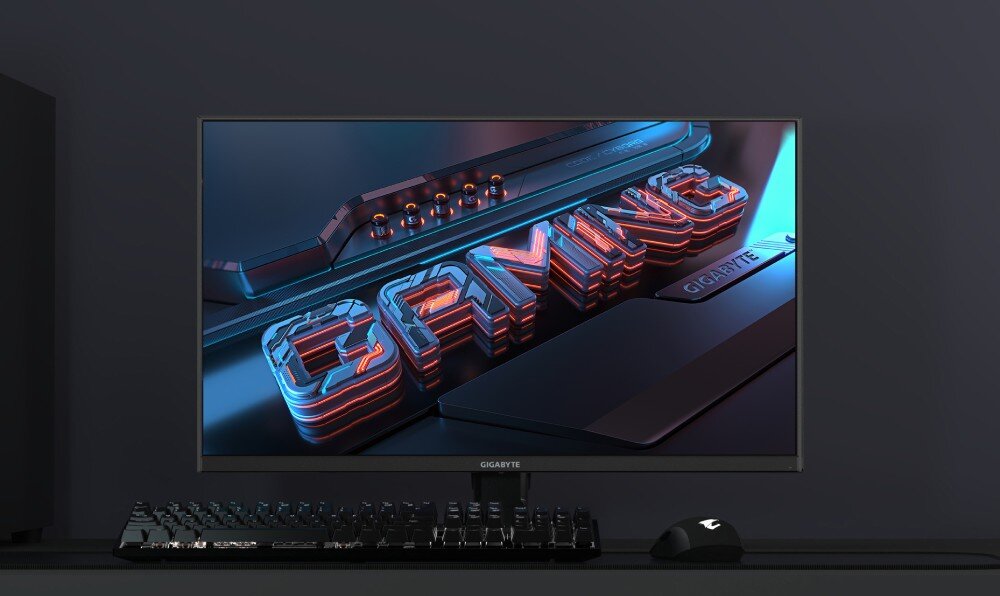 Monitor GIGABYTE GS27QXA Ekran monitora wyświetlający trójwymiarowy napis 'GAMING' w kolorach niebieskim i czerwonym, ustawiony na biurku obok klawiatury i myszy, opis