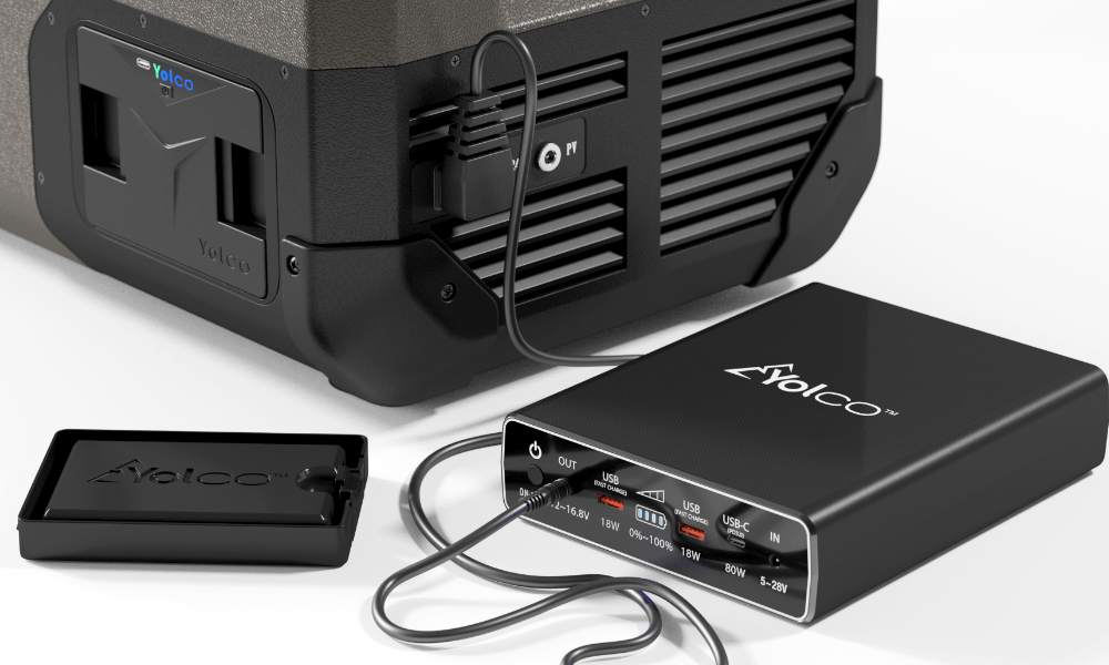 PRZENOŚNA LODÓWKA KOMPRESOROWA YOLCO FCX18 CREAM Lodówka podłączona jest do zewnętrznego modułu zasilania z portami USB i USB‑C, a obok widoczna jest zdejmowana osłona baterii. Kable łączą urządzenia, tworząc zestaw do zasilania i ładowania zasilanie 12V 24V 230V bateria Yolco panel fotowoltaiczny USB ładowanie port USB panel sterowania łatwa obsługa lodówka mobilna uchwyt transportowy