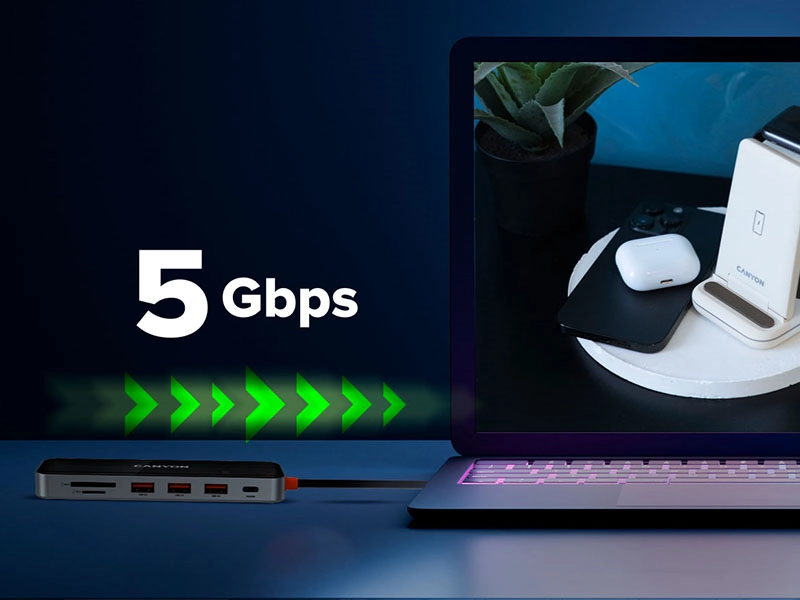 Na obrazie widać laptopa z podłączonym hubem USB-C marki Canyon, obok którego znajduje się napis \'5 Gbps\'. Na ekranie laptopa widoczny jest stojak z urządzeniami elektronicznymi oraz rośliną.