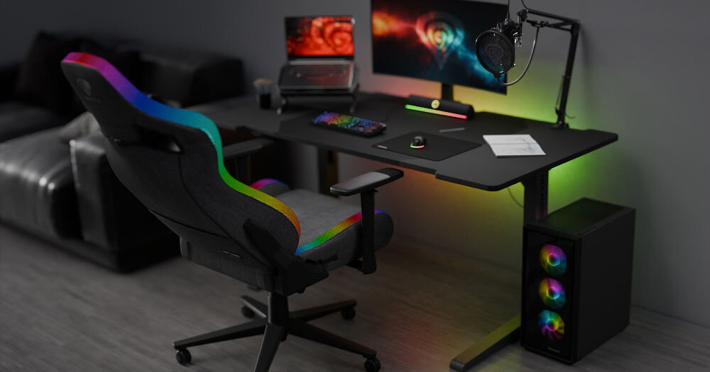 Fotel GENESIS Trit 660 RGB Szary fotel z włączonym kolorowym podświetleniem wokół siedziska i oparcia stojący przy biurku z komputerem gamingowym poduszka lędźwiona zagłówkowa pianka zapamiętująca kształt wsparcie komfort poszerzone siedzisko oparcie