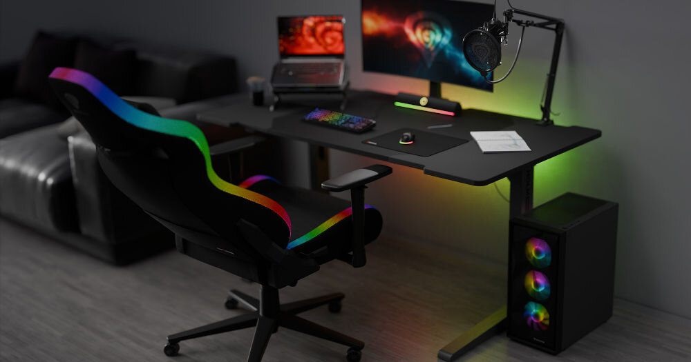 Fotel GENESIS Trit 660 RGB Czarny fotel z włączonym kolorowym podświetleniem wokół siedziska i oparcia stojący przy biurku z komputerem gamingowym poduszka lędźwiona zagłówkowa pianka zapamiętująca kształt wsparcie komfort poszerzone siedzisko oparcie