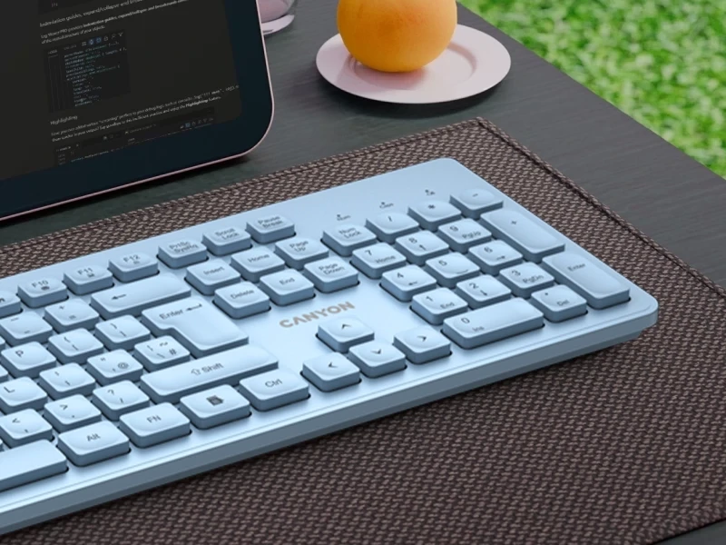 Na biurku znajduje się niebieska klawiatura z napisem \'CANYON\'. Obok klawiatury leży tablet z wyświetlonym kodem oraz talerzyk z owocem.