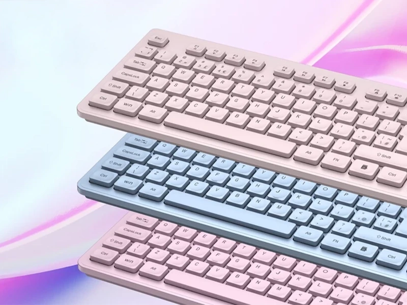 Na obrazie widoczne są trzy bezprzewodowe klawiatury w pastelowych kolorach: dwie różowe i jedna niebieska, ułożone jedna nad drugą na tle w odcieniach różu i fioletu.