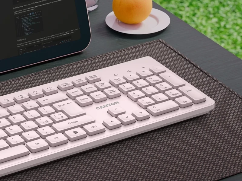 Beżowa klawiatura bezprzewodowa z logo \'CANYON\' na biurku obok laptopa i talerzyka z owocem. Klawiatura ma standardowy układ klawiszy z widocznymi oznaczeniami liter i cyfr.