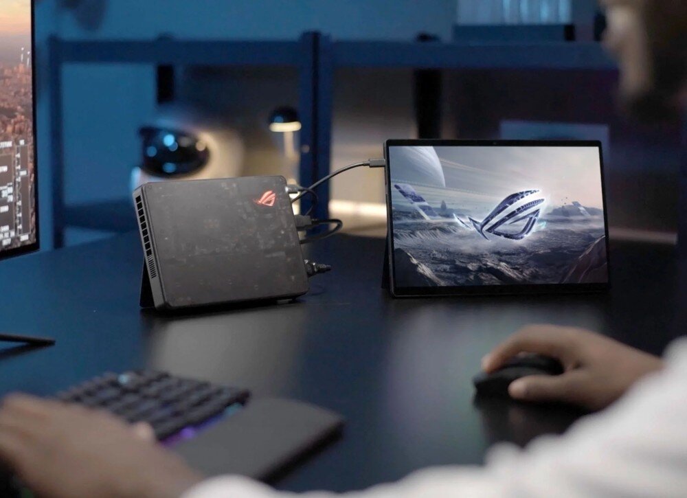 Karta graficzna ASUS ROG XG Mobile GC34R GeForce RTX 5070 Ti 12GB zewnętrzna stacja graficzna z podświetlonym na czerwono logo stoi na biurku obok tabletu, przy którym osoba posługuje się myszką i klawiaturą