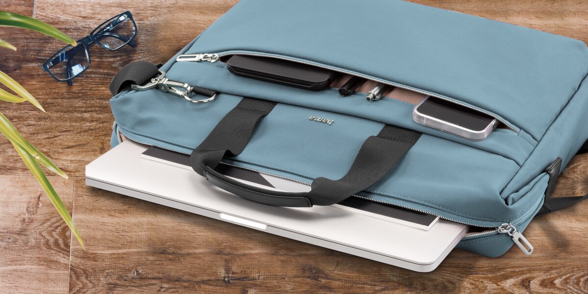 Torba na laptopa HAMA Ultimate 13.3 - 14.1 cala Niebieski Torba na laptopa Hama leżąca na drewnianym blacie z otwartą przegrodą, z której wysunięty jest biały laptop.