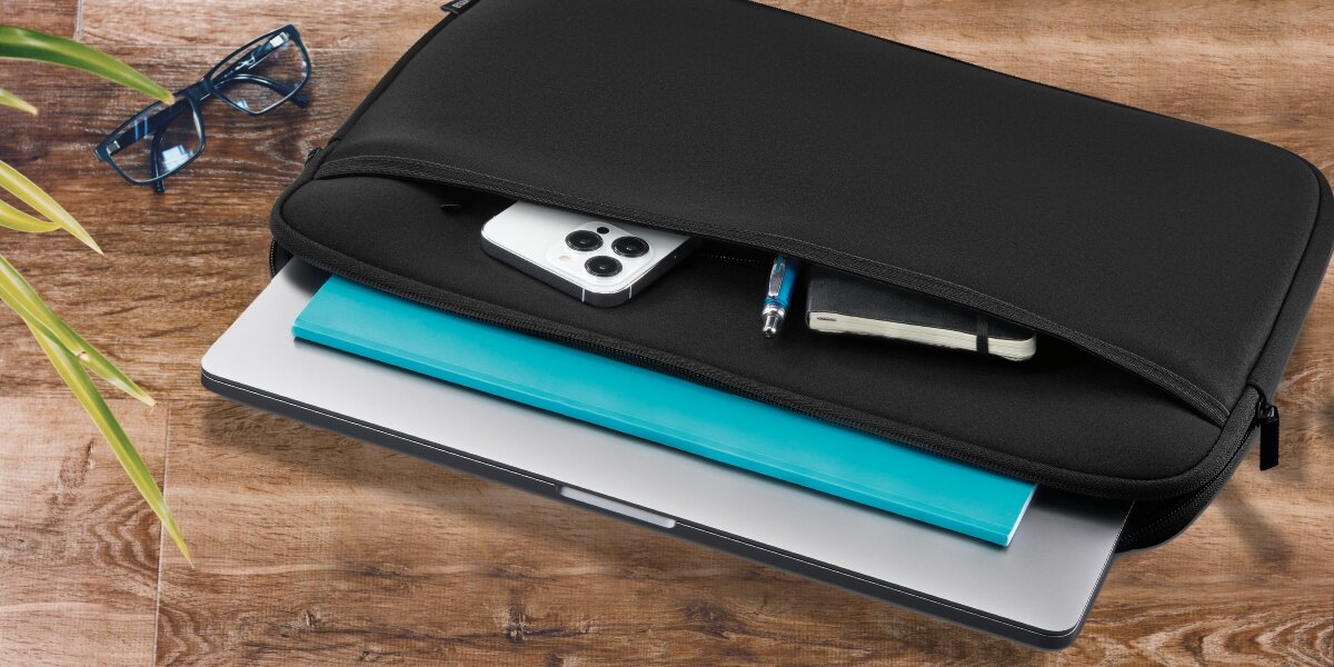 Torba na laptopa HAMA Neoprynowy 14.1-15.6 Czarny pokrowiec na laptopa Hama z wysuniętym smartfonem, długopisem i notesem z przedniej kieszeni, leżący obok okularów na drewnianym blacie. 