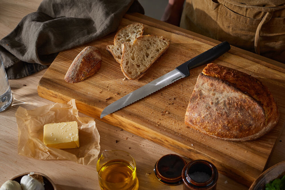 Nóż FISKARS Hard Edge 1054945 nóż z czarną rączką leży na desce obok leży chleb Ostrze z wysokiej jakości japońskiej stali nierdzewnej precyzja efektywność trwałość dynamiczne cięcie z dużą siłą subtelna precyzja w codziennym gotowaniu przy wymagających technikach kulinarnych