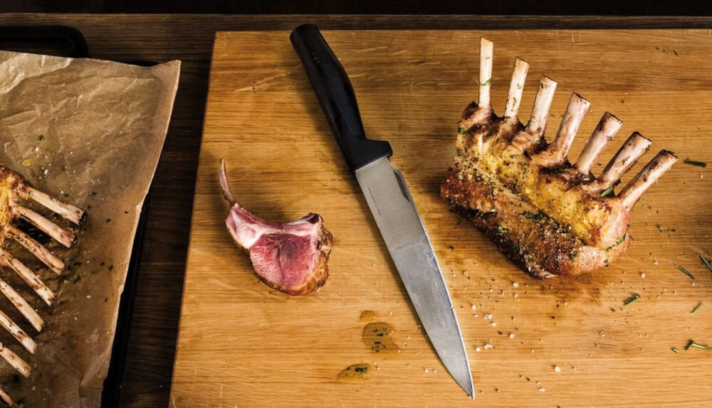 Nóż FISKARS Hard Edge 1051760 deska na desce znajduje się nóż z czarną rękojeścią obok leży mięso dla każdego Ostrze o długości 21,6 cm całkowitej długości 35,1 cm porcjowanie mięsa waży jedynie 113 g