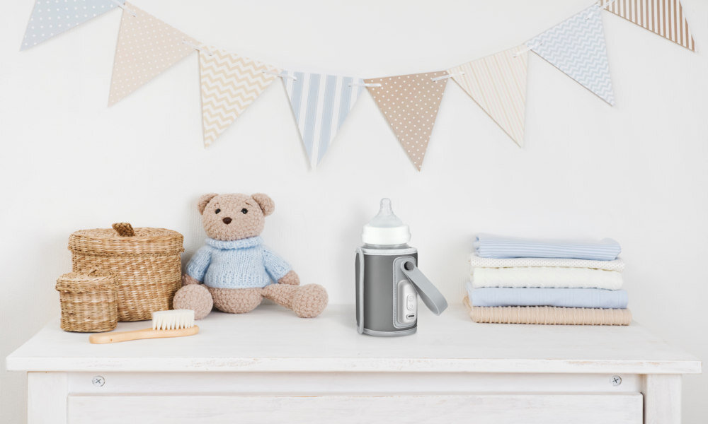 Na białej komodzie znajduje się srebrny podgrzewacz do butelek oraz stos złożonych ręczników. W tle widać pluszowego misia i koszyki wiklinowe.