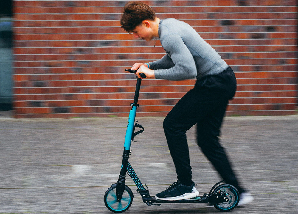 Hulajnoga NILS EXTREME HM124T Turkusowy droga miasto osoba jedzie na hulajnodze po drodze w tle budynek z czerwonej cegły pasja hobby komfortowa jazda mobilność