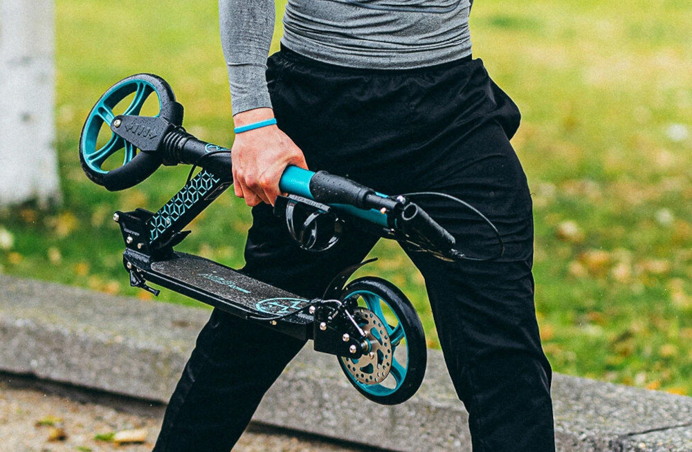 Hulajnoga NILS EXTREME HM124T Turkusowy park osoba idzie po drodze ze złożoną hulajnogą w dłoni w tle trawnik mechanizm składania lekka konstrukcja waga 5,6 kg składana konstrukcja wraz ze składanymi uchwytami po złożeniu zajmuje niewiele miejsca