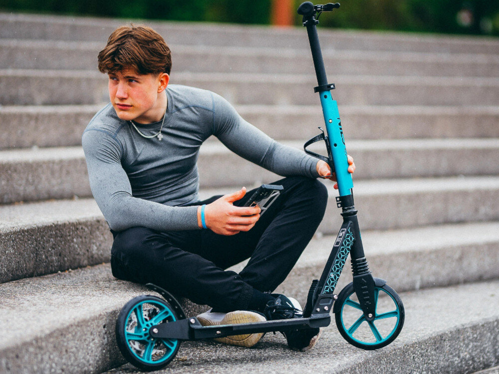 Hulajnoga NILS EXTREME HM124T Turkusowy betonowe schody osoba siedzi na schodach patrz za siebie przed sobą ma hulajnogę łączenie kierownicy z deckiem białe tło z aluminium spełnia wymagania normy EN71-1:2014 bezpieczeństwo oraz jakość wykonania maksymalne obciążenie do 100 kg odporna na codzienne użytkowanie łączy stabilność z komfortem przenoszenia