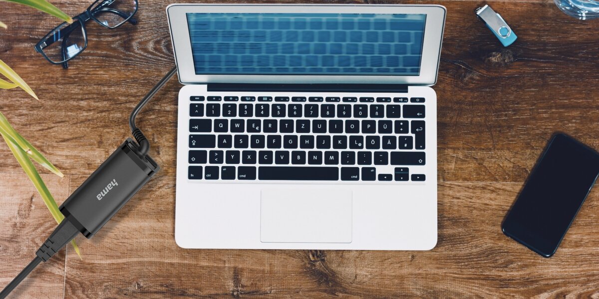 Czarna ładowarka Hama USB-C 45W podłączona do otwartego laptopa (prawdopodobnie MacBooka) leżącego na drewnianym biurku. Obok leży smartfon, okulary i pendrive.