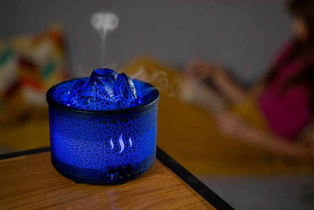 Aromatyzer GÖTZE & JENSEN AH202 Volcano ozdoba wnętrza design black crack dekoracja do salonu - Dyfuzor zapachowy w kształcie wulkanu, świecący na intensywny niebieski kolor z efektem spękanej powierzchni, emituje delikatną mgiełkę. W tle siedzi zamazana sylwetka kobiety