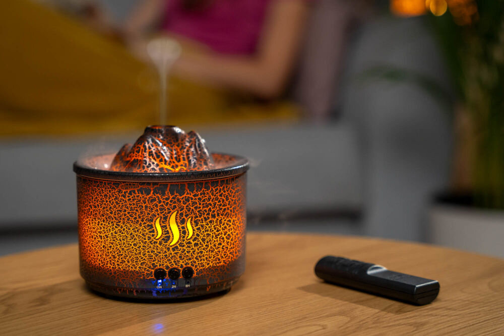 Aromatyzer GÖTZE & JENSEN AH202 Volcano podświetlenie LED pomarańcz niebieski nastrój w domu - Dyfuzor zapachowy w kształcie wulkanu z efektem spękanej lawy, intensywnie podświetlony na ciepły, pomarańczowy kolor (imitujący ogień). Z wierzchu unosi się para. Obok dyfuzora leży czarny pilot zdalnego sterowania