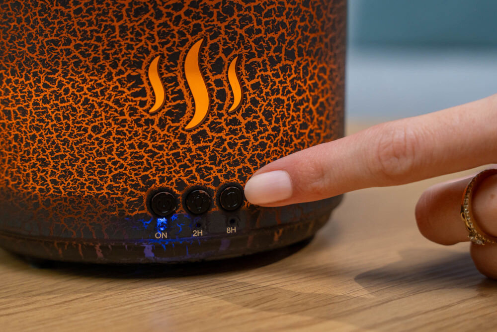 Dyfuzor zapachowy GÖTZE & JENSEN AH202 Volcano automatyczne wyłączanie bezpieczeństwo użytkowania funkcje ochronne Zbliżenie na spód dyfuzora zapachowego. Palec użytkownika naciska jeden z przycisków timera (ON, 2H, 8H). Świeci się niebieska dioda