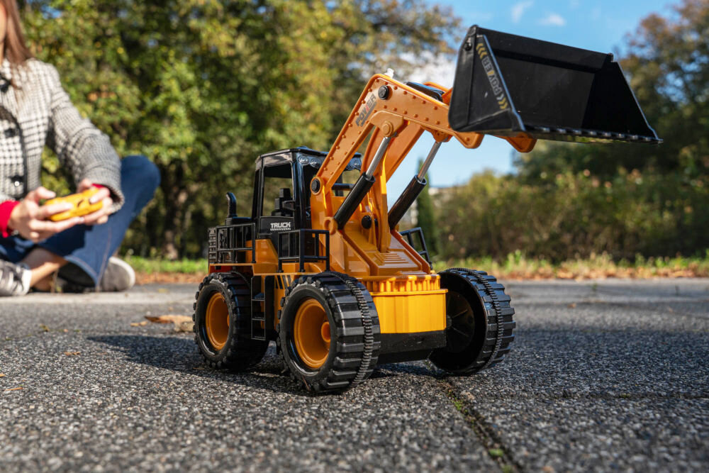 ŁADOWARKA RC S-LINE RCC204 Zabawkowa, pomarańczowo-czarna ładowarka kołowa na środku okrągłego, fioletowego dywanu w jasnym pokoju zabaw. Wokół zabawki leżą klocki, piłka i małe samochodziki. W tle widoczny jest pluszowy krokodyl i indiański namiot (tipi).