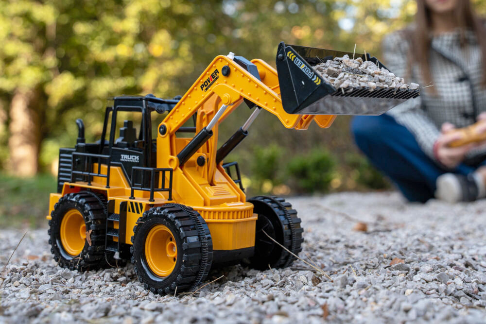 ŁADOWARKA RC S-LINE RCC204 Zabawkowa ładowarka kołowa ustawiona na kolorowym dywanie z motywem zwierząt w pokoju dziecięcym. W tle widoczne jest białe łóżeczko z pościelą w niebieskie dinozaury i biały regał z książkami i zabawkami.