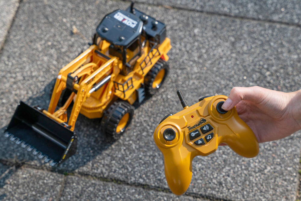 ŁADOWARKA RC S-LINE RCC204 Zabawkowa ładowarka kołowa na drewnianej podłodze w pokoju dziecięcym. Jest to ujęcie z niskiej perspektywy, ukazujące zabawkę w otoczeniu porozrzucanych klocków, pluszowych misiów i innych zabawek. W tle duże okno i regał.