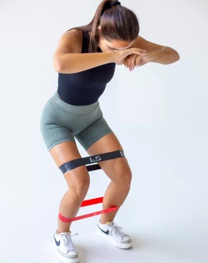 Osoba w sportowym stroju wykonuje ćwiczenie z użyciem dwóch gum Mini Power Band, jednej czarnej i jednej czerwonej, umieszczonych wokół ud i łydek.