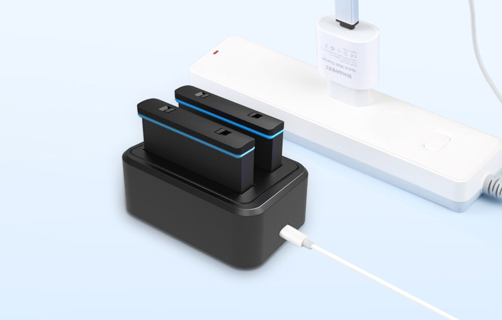 Na obrazie widoczna jest czarna ładowarka dwukanałowa z dwoma bateriami, podłączona do białej listwy zasilającej. W tle znajduje się biały adapter z logo \'Huawei\'.