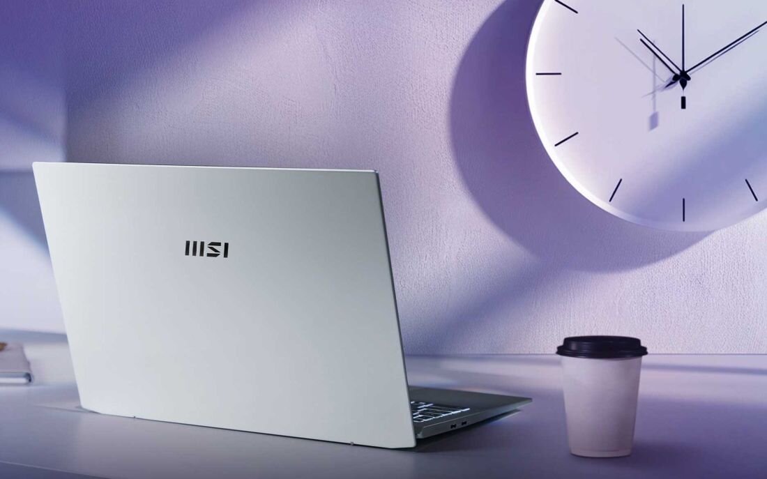 Laptop MSI Prestige A16 AI+ w kolorze srebrnym na jasnym biurku, w tle duży, minimalistyczny zegar ścienny i kubek z pokrywką – ilustracja długiego czasu pracy na baterii oraz eleganckiego designu. - Wytrzymała bateria 82 Wh