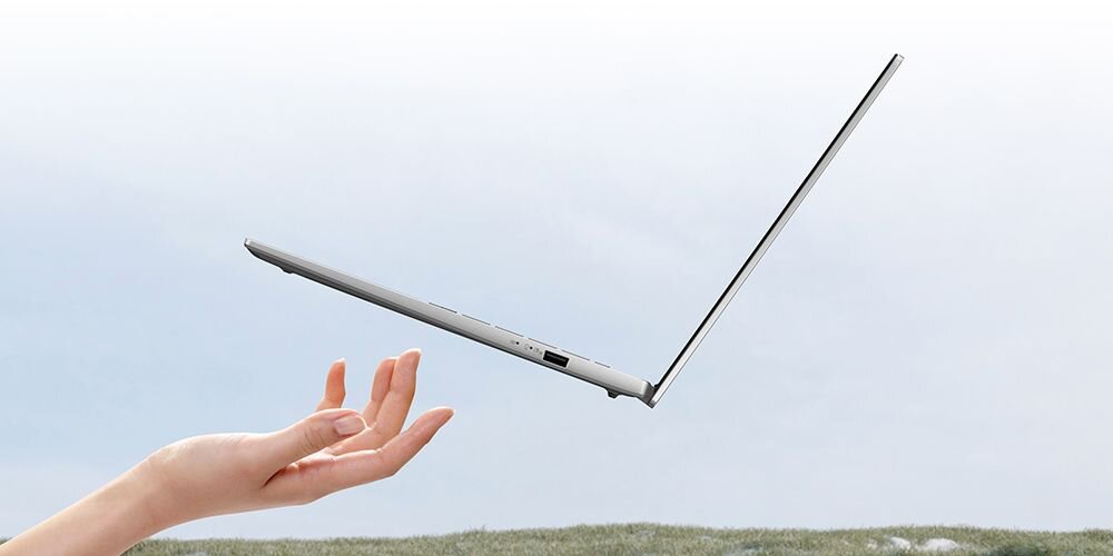 Lekki, smukły laptop ASUS VivoBook S 14 w kolorze srebrnym unoszący się nad dłonią na tle nieba i trawy, podkreślający mobilność i niewielką wagę urządzenia. - Smukły design