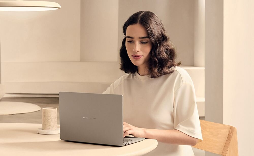 Kobieta w jasnej bluzce siedzi przy stole i używa laptopa ASUS Zenbook w kolorze szarym, podkreślając energooszczędną platformę Snapdragon X i szybkie ładowanie USB-C PD - Bateria 70 Wh
