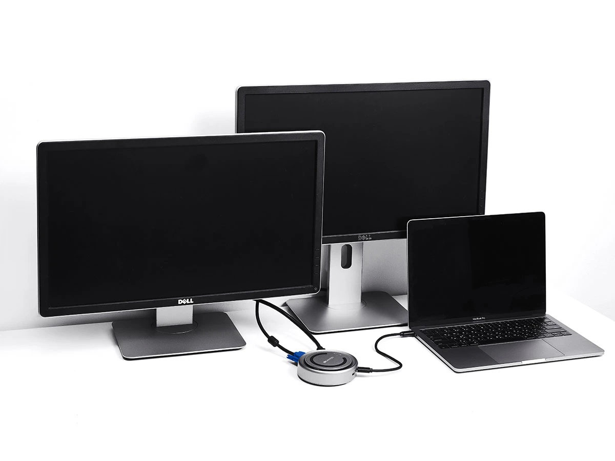 Na białym biurku znajdują się dwa monitory i laptop, wszystkie z wyłączonymi ekranami. Obok laptopa leży okrągły hub USB-C z podłączonymi kablami.