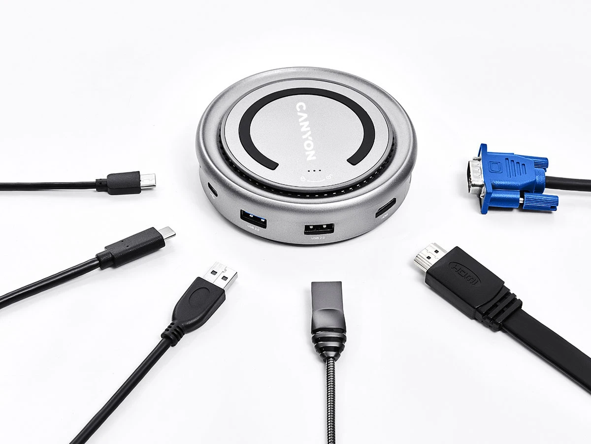 Na obrazie widoczny jest okrągły, srebrny hub z napisem \'CANYON\' na górze, otoczony różnymi kablami, w tym USB, HDMI i VGA. Hub posiada kilka portów, w tym USB 3.0 i USB 2.0.