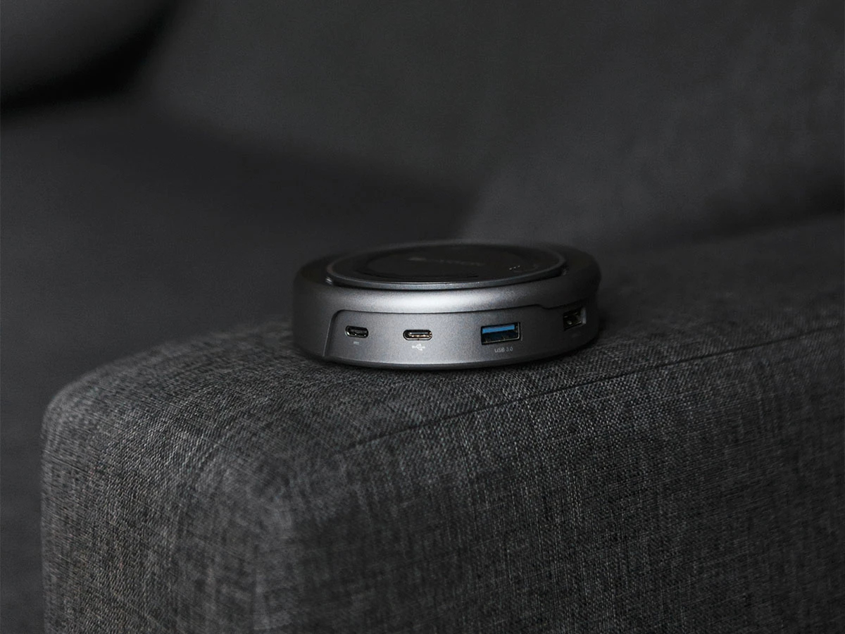 Na ciemnoszarej kanapie leży okrągły, szary hub USB z kilkoma portami, w tym USB-C i USB 3.0. Produkt ma gładką powierzchnię i kompaktowy design.
