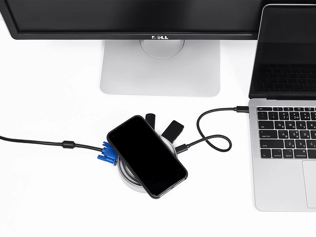 Na białym biurku znajduje się monitor z logo \'Dell\', laptop oraz smartfon leżący na okrągłym urządzeniu z kilkoma podłączonymi kablami. Urządzenie jest połączone z laptopem za pomocą kabla USB-C.