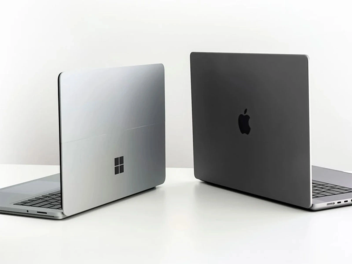 Na białym stole znajdują się dwa laptopy, jeden z logo w kształcie okna, a drugi z logo w kształcie jabłka. Oba urządzenia są otwarte i ustawione tyłem do widza.
