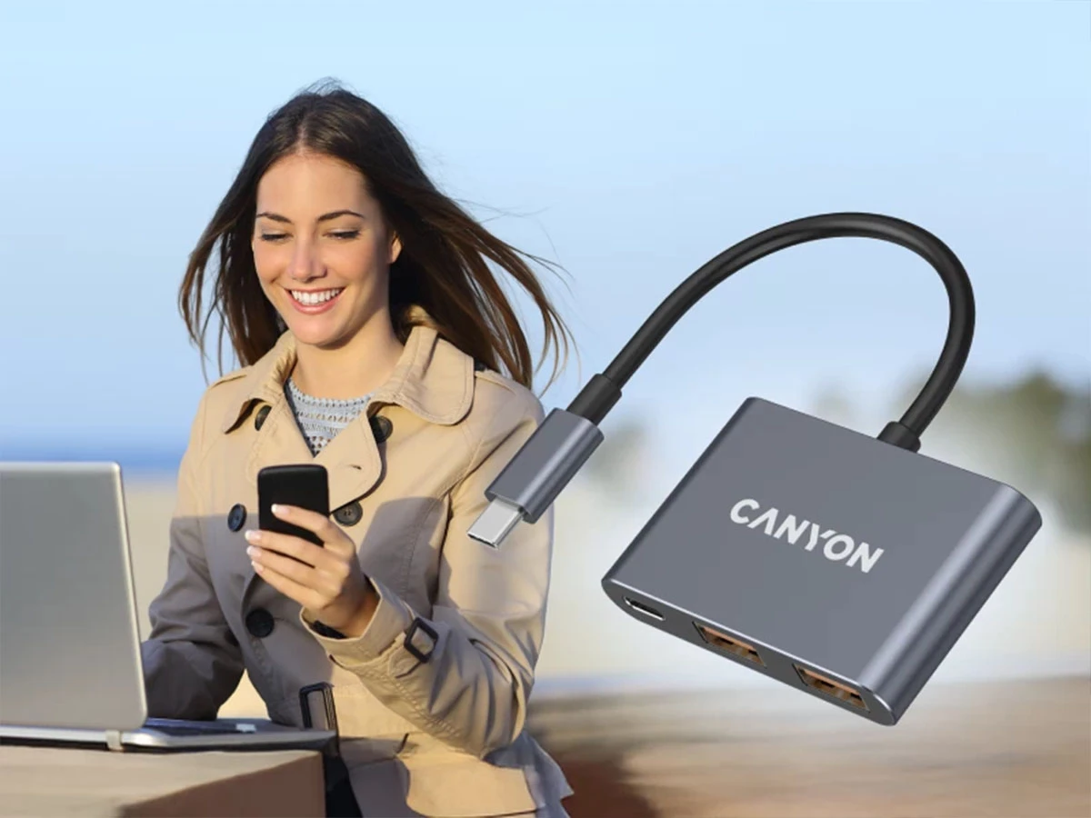 Kobieta siedzi przy laptopie i trzyma telefon, obok niej unosi się ciemnoszary hub USB-C z napisem \'CANYON\'.
