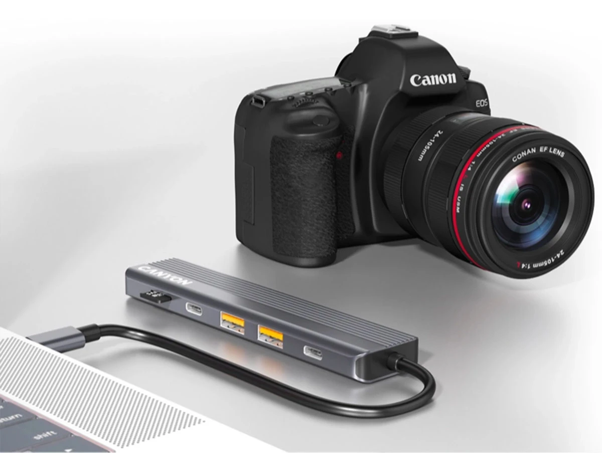Na obrazie widoczny jest aparat fotograficzny z obiektywem oraz szary hub USB-C z logo \'CANYON\', podłączony do laptopa. Hub posiada kilka portów, w tym dwa żółte i jeden czarny.
