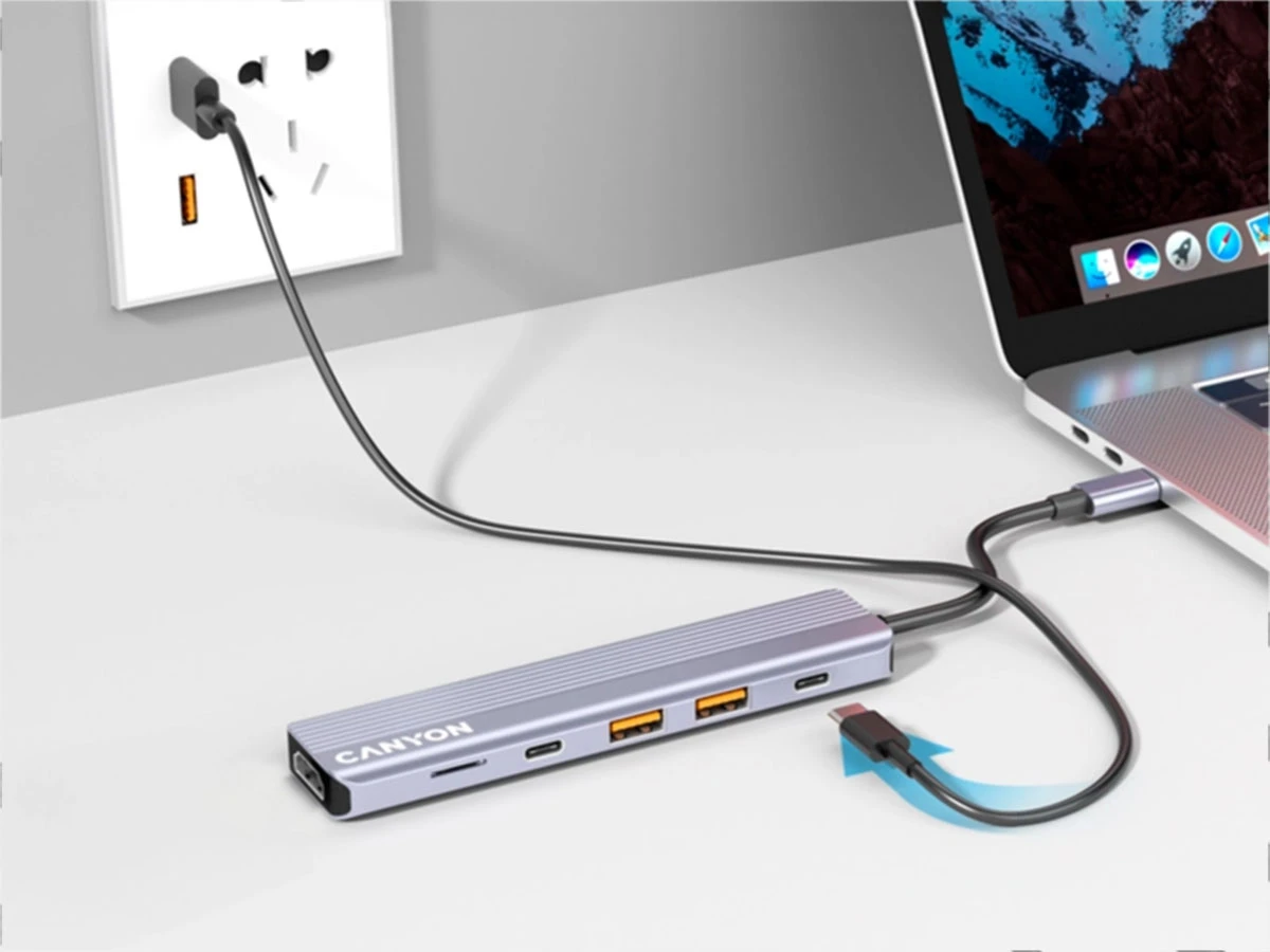 Na biurku znajduje się szary hub USB-C z logo \'CANYON\', podłączony do laptopa i gniazdka elektrycznego. Hub posiada kilka portów, w tym USB i HDMI.
