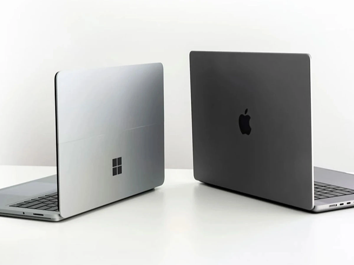 Na białym tle znajdują się dwa laptopy, jeden z logo w kształcie okna, a drugi z logo w kształcie jabłka. Oba urządzenia są otwarte i ustawione tyłem do siebie.
