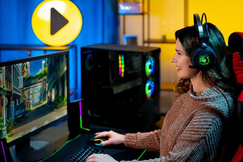 Komputer MAD DOG ARYA-I04DR16 Kobieta w słuchawkach z mikrofonem siedzi przy biurku i gra na komputerze gamingowym z podświetlanymi wentylatorami. Na monitorze widoczna animowana scena przedstawiająca ulicę, przeznaczenie, testy, podzespoły