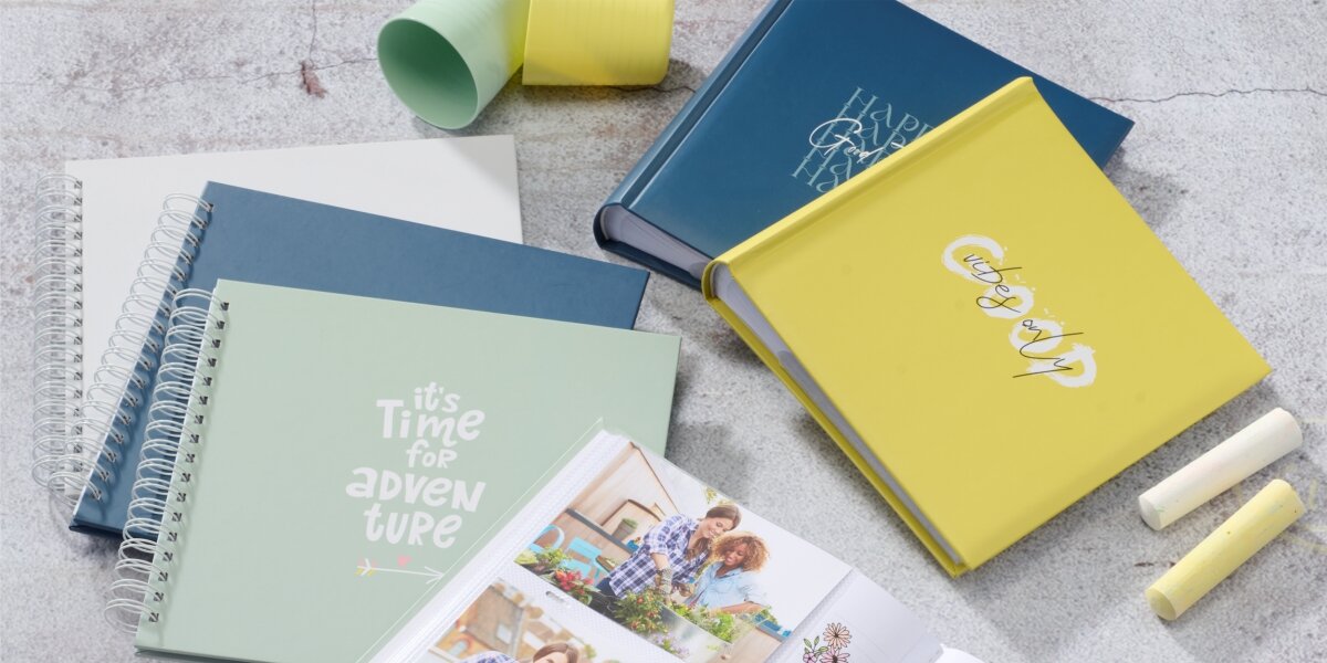 Na obrazie widoczne są różne albumy fotograficzne w pastelowych kolorach, w tym zielony z napisem \'it\'s Time for adventure\' oraz żółty z napisem \'Good Things Happen\'. Obok albumów leżą kredki i rozwinięty album z fotografiami.