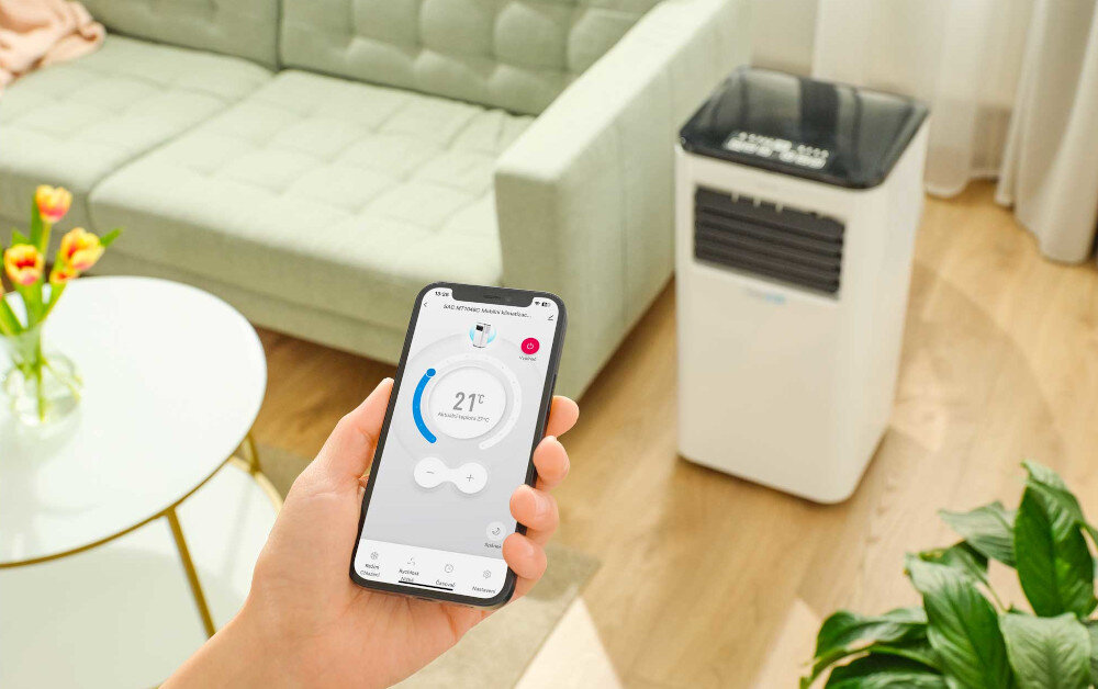 Osoba trzyma smartfon z aplikacją pokazującą temperaturę 21°C, w tle widać biały klimatyzator przenośny w jasnym pokoju z zieloną sofą i rośliną.