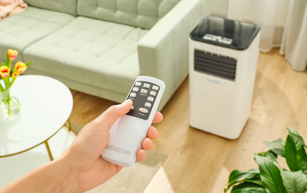 Osoba trzyma pilot z napisem \'SENCOR\', w tle widoczny jest biały klimatyzator przenośny w jasnym wnętrzu z zieloną sofą i roślinami.