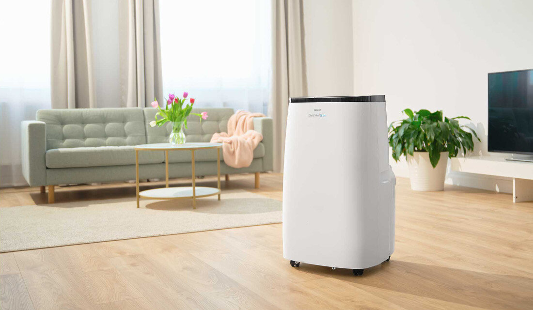 Biały klimatyzator stoi na drewnianej podłodze w jasnym salonie z sofą, stolikiem i rośliną w tle. Na urządzeniu widoczny jest napis \'SENCOR SAC MT1803CH\'.