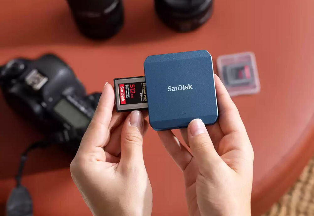 Czytnik kart SANDISK PRO-CINEMA CFexpress Typ B prezentacja czytnika podczas wkładania karty czytnik trzymany w rękach w tle aparat i obiektywy iPhone 15 Pro | Pro Max – rejestrowanie ProRes, montaż MagSafe, nagrywanie 4K