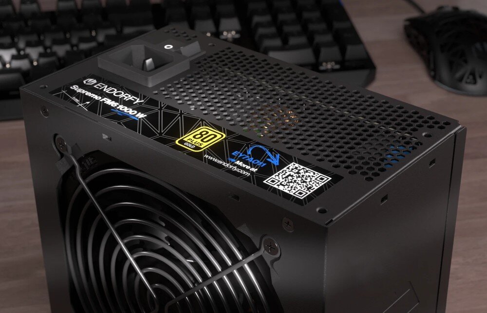 Komputer MAD DOG ENDORFY700AIR-A02DB32 Zbliżenie na zasilacz ustawiony na biurku. Widoczna jest naklejka informacyjna z oznaczeniem modelu, certyfikatem 80 PLUS Gold, kodem QR. W tle widać fragment klawiatury i myszy komputerowej, zasilacz, moc, sprawność, kondensatory