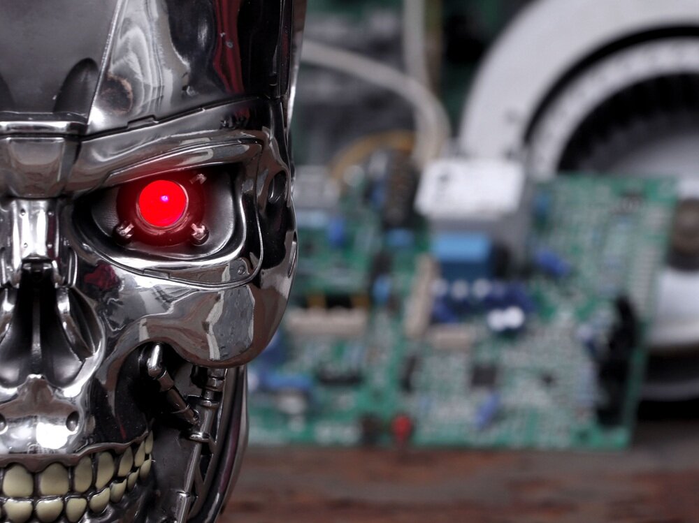 Metaliczna głowa robota z czerwonym, świecącym okiem na tle rozmytych elementów elektronicznych.