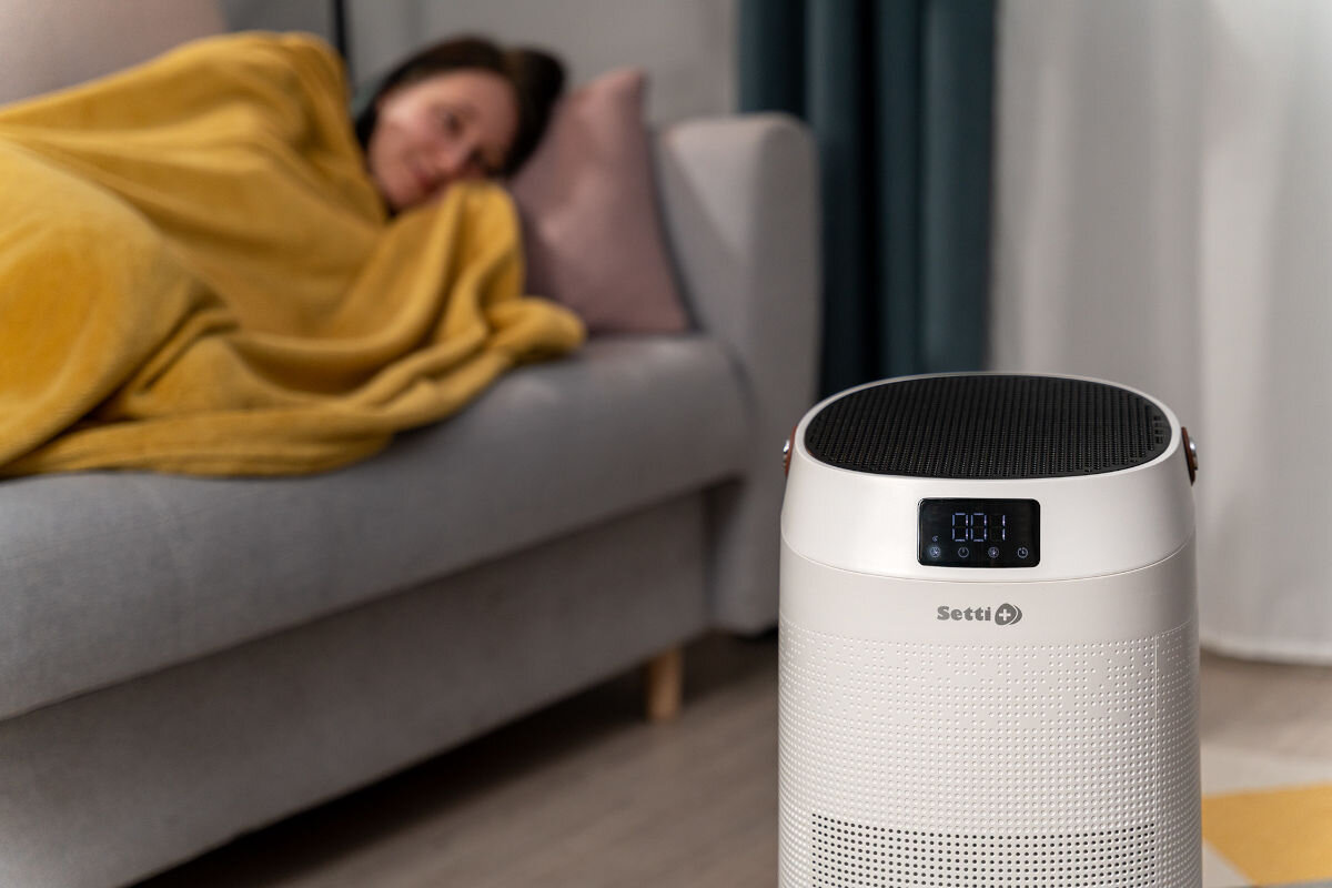 Oczyszczacz powietrza SETTI+ AP650W Wi‑Fi Biały oczyszczacz powietrza Setti+ z wyraźnym wskaźnikiem jakości powietrza 001 na wyświetlaczu. Na drugim planie, zamazana sylwetka kobiety owiniętej żółtym kocem, odpoczywającej na kanapie.