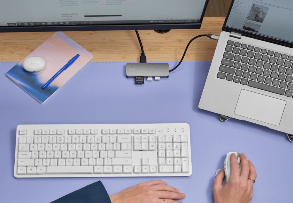 Hub TRUST Dalyx 25672 Widok z góry na dłonie osoby korzystającej z białej myszy i klawiatury przy biurku z laptopem podłączonym do wieloportowego adaptera z włożoną kartą pamięci