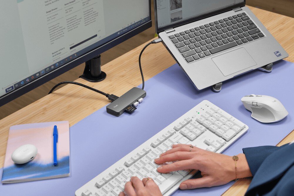 Hub TRUST Dalyx 25672 Stanowisko pracy z laptopem na podstawce podłączonym do szarego huba obsługującego monitor oraz białą klawiaturę i mysz używane przez osobę przy biurku